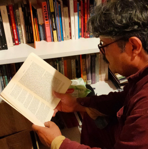 a man with grey hair and a bright coloured shirt on is turned side on to the camera, he is reading from a book that is only text. In the background of the image is a bookshelf full of books