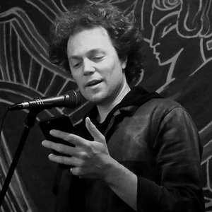 Male Poet reading from his phone into a microphone, there is a dark patterned wall behind him